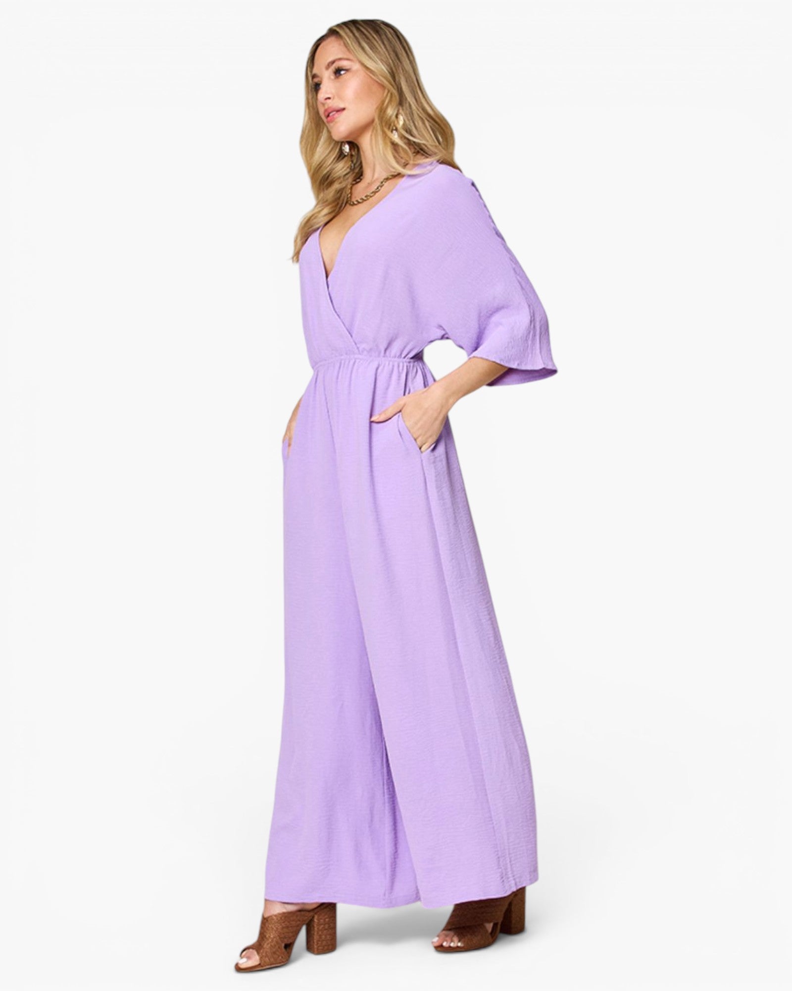 A woman modeling a lavender purple maxi jumpsuit with wide three-quarter sleeves, a wrap-style V-neckline, and side pockets, paired with brown heeled sandals against a plain white studio background.