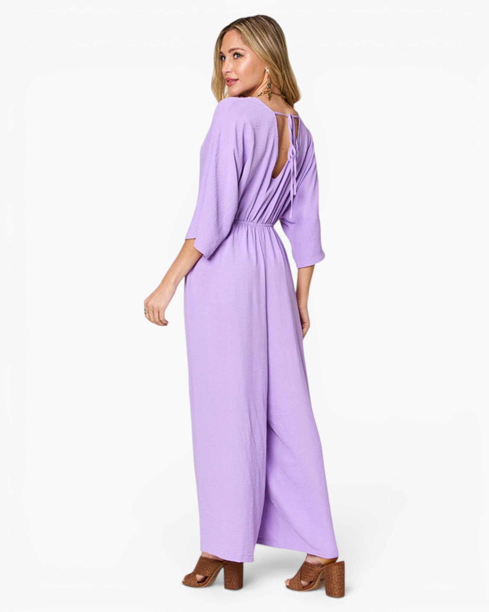 A woman modeling a lavender purple jumpsuit with three-quarter sleeves, a wrap-style waist, and an open back, shown against a plain white studio background.