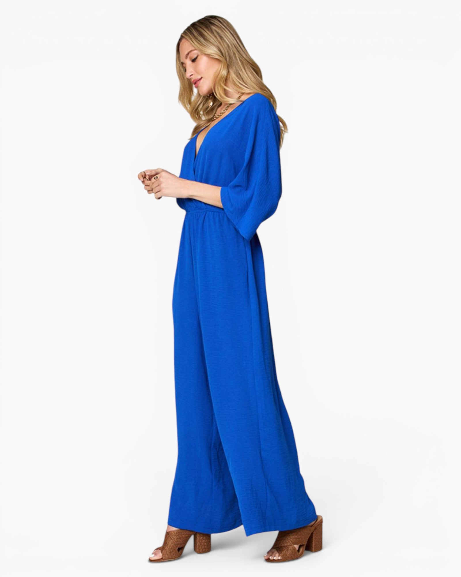 A woman modeling a bright royal blue jumpsuit with three-quarter sleeves and wide legs, paired with brown heeled sandals, against a plain white studio background.