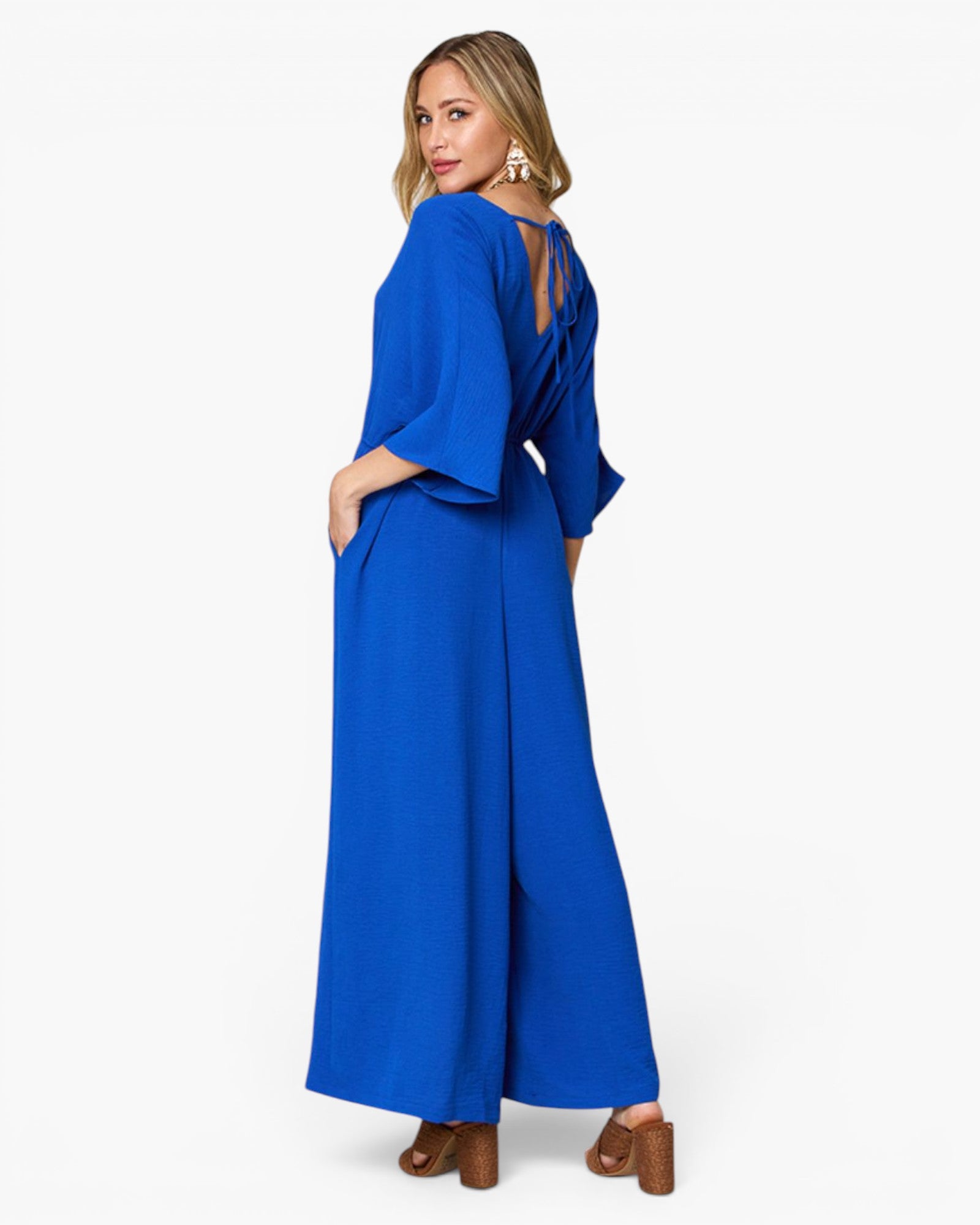 A woman modeling a bright royal blue wide-leg jumpsuit from the back, featuring three-quarter bell sleeves, side pockets, and an open back design, set against a plain white studio background.