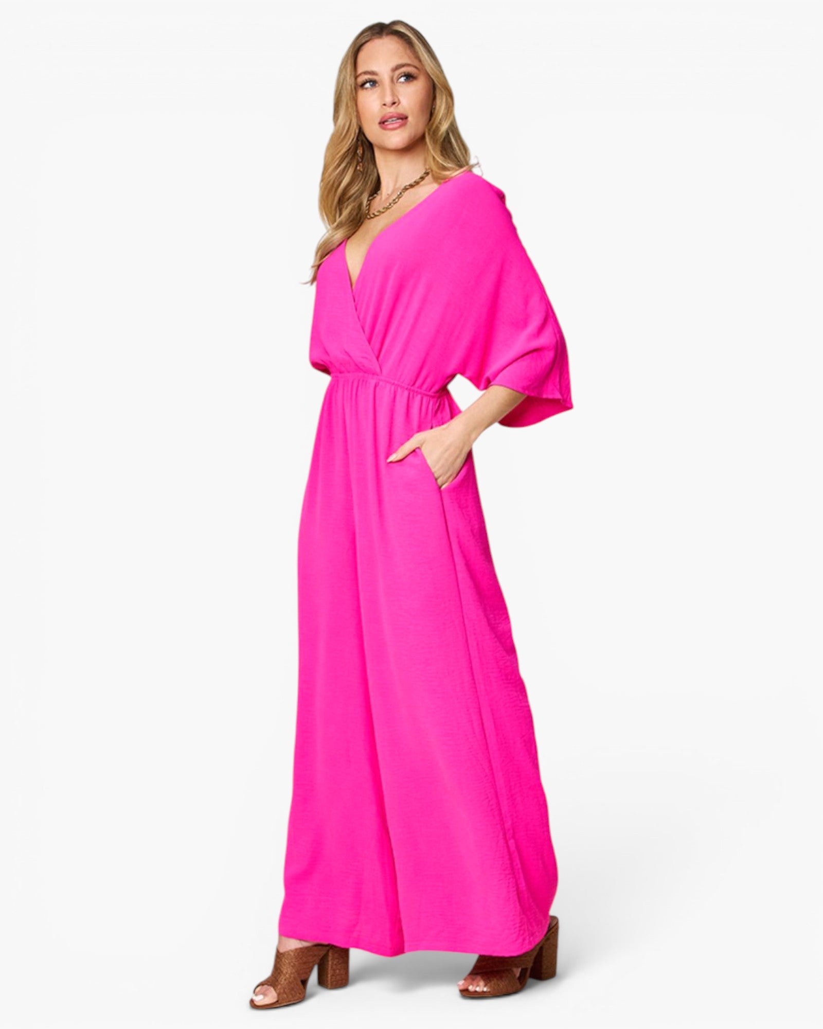 A woman modeling a bright hot pink maxi dress with a deep V-neckline, draped three-quarter sleeves, and side pockets, paired with brown heeled sandals against a plain white studio background.