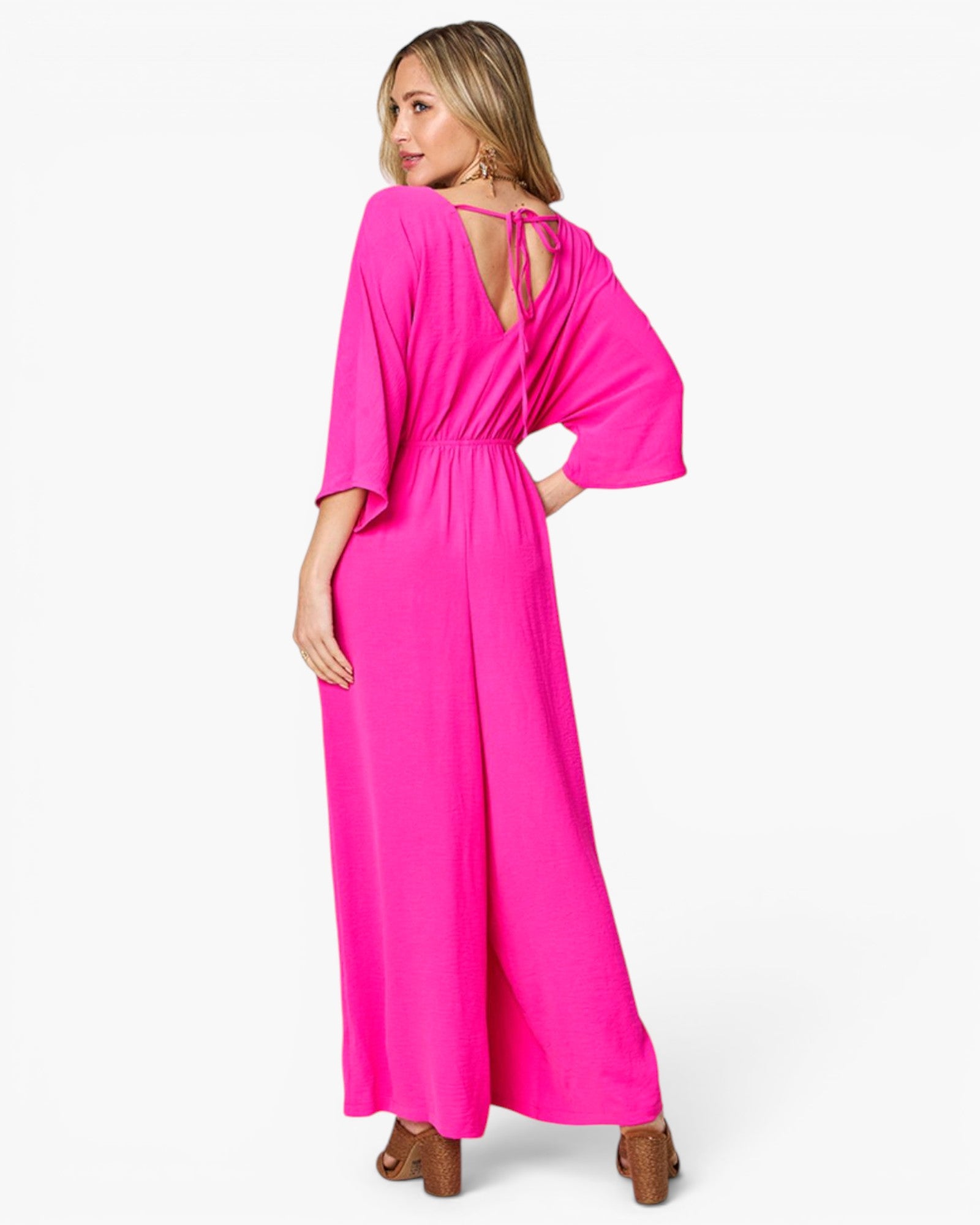 A woman modeling a bright pink maxi jumpsuit from the back, featuring a tie-back neckline, three-quarter flared sleeves, and wide legs, set against a plain white studio background.