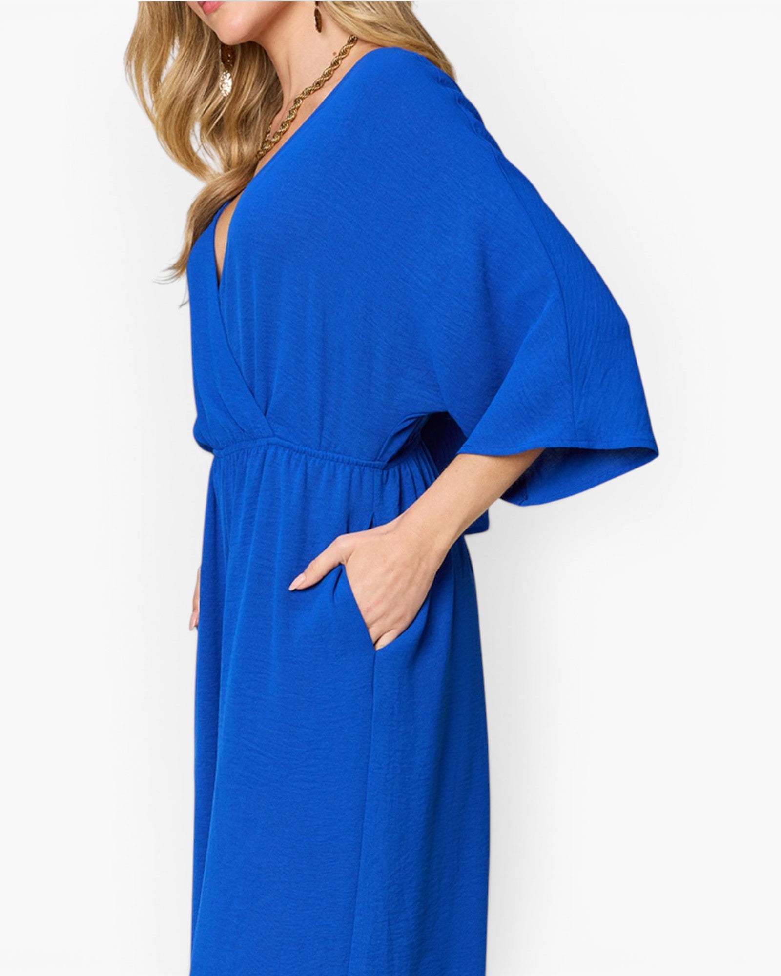 A woman wearing a bright royal blue wrap-style maxi dress with wide flutter sleeves and side pockets, shown from the side against a plain white background.