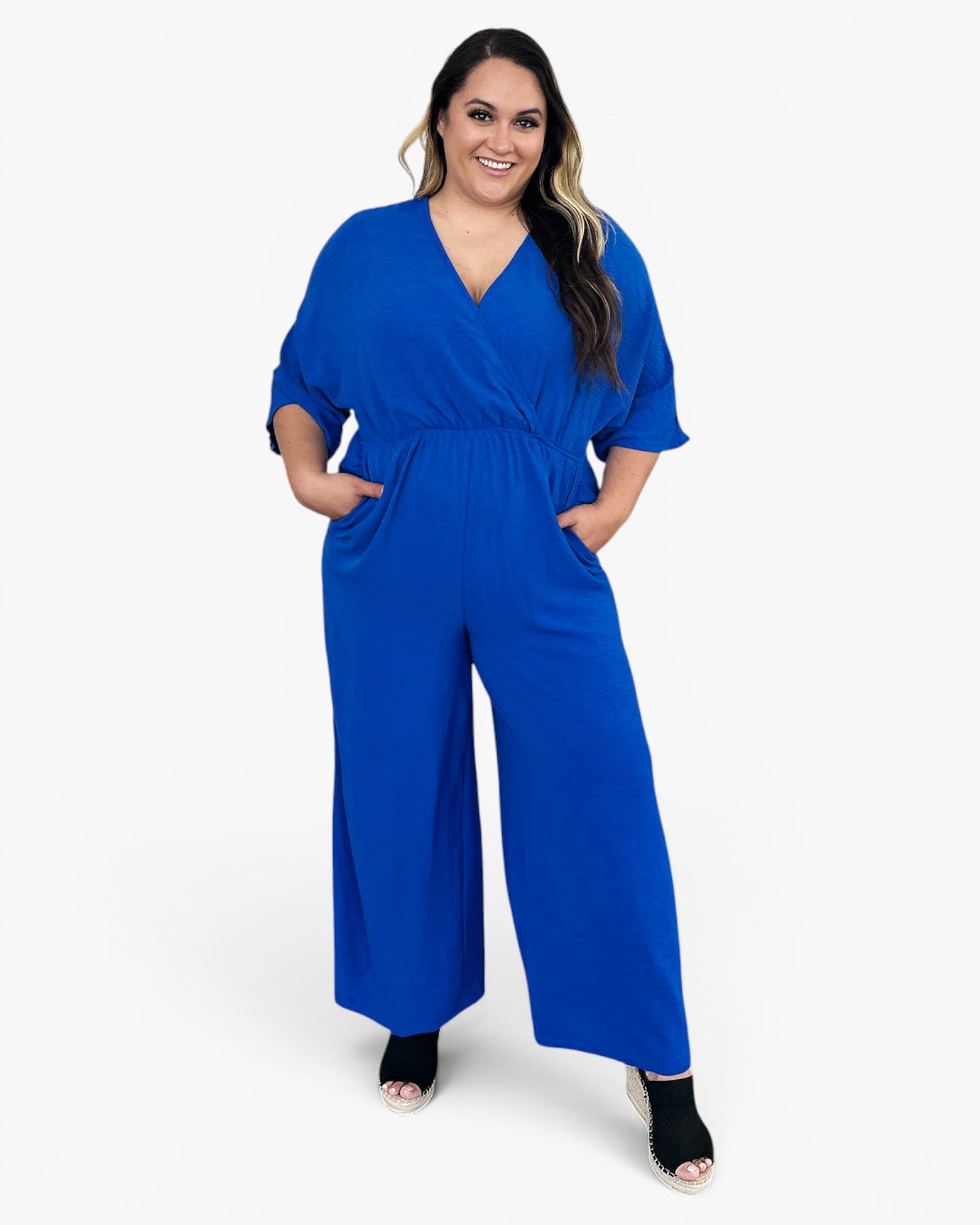 A woman modeling a bright royal blue jumpsuit with short sleeves, a V-neckline, and wide legs, standing against a plain white studio background.