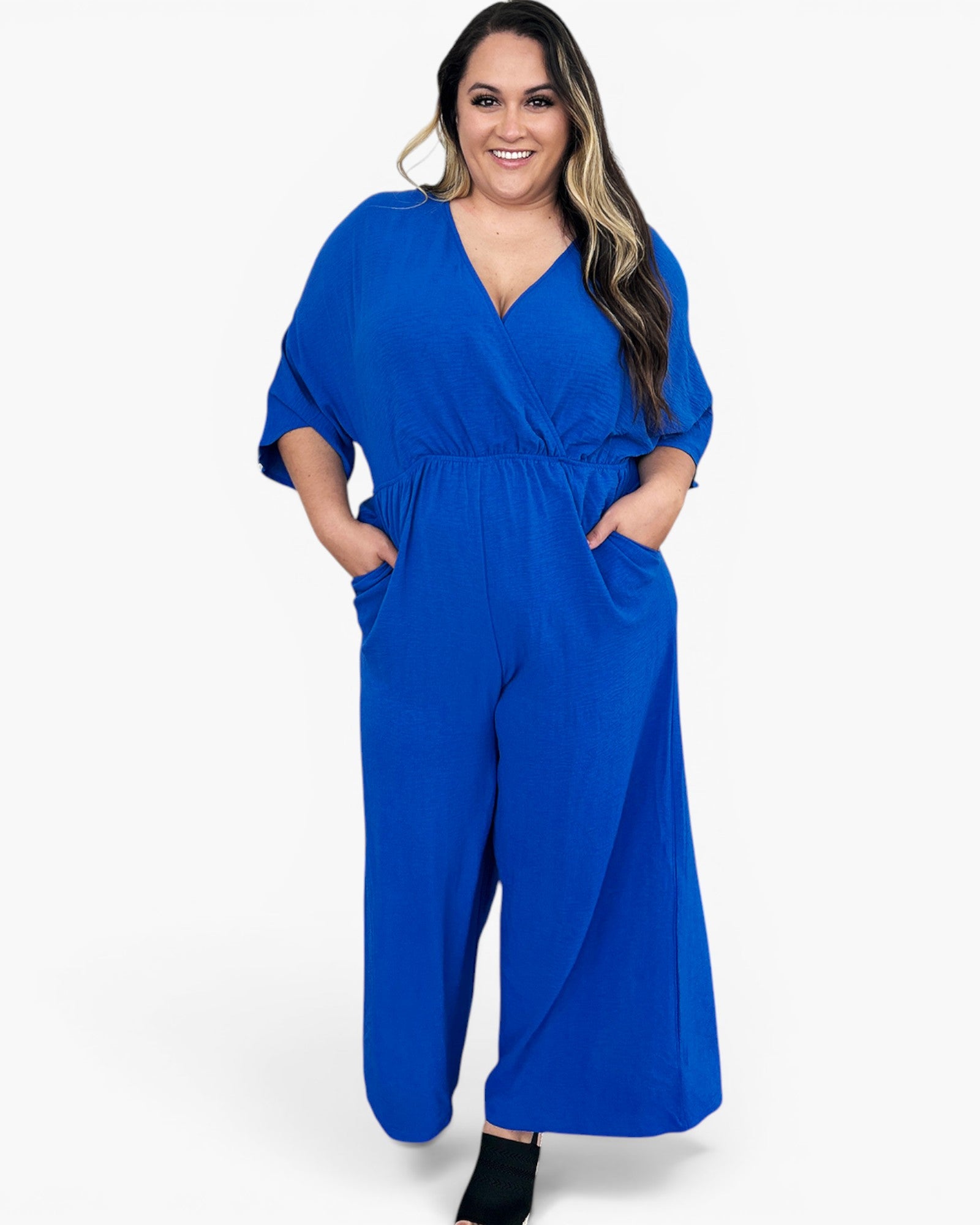 A woman modeling a bright royal blue wide-leg jumpsuit with a V-neck and elbow-length sleeves, standing against a plain light gray studio background.