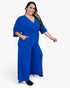 A woman modeling a bright royal blue wrap-style jumpsuit with wide legs, elbow-length sleeves, and side pockets, photographed against a clean white studio background.