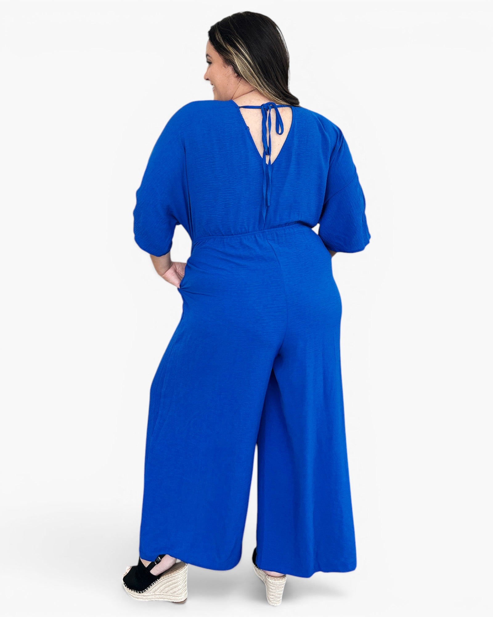 A woman wearing a bright royal blue wide-leg jumpsuit, shown from the back against a plain white studio background.