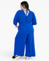 A woman wearing a bright royal blue wide-leg jumpsuit, shown from the back against a plain white studio background.