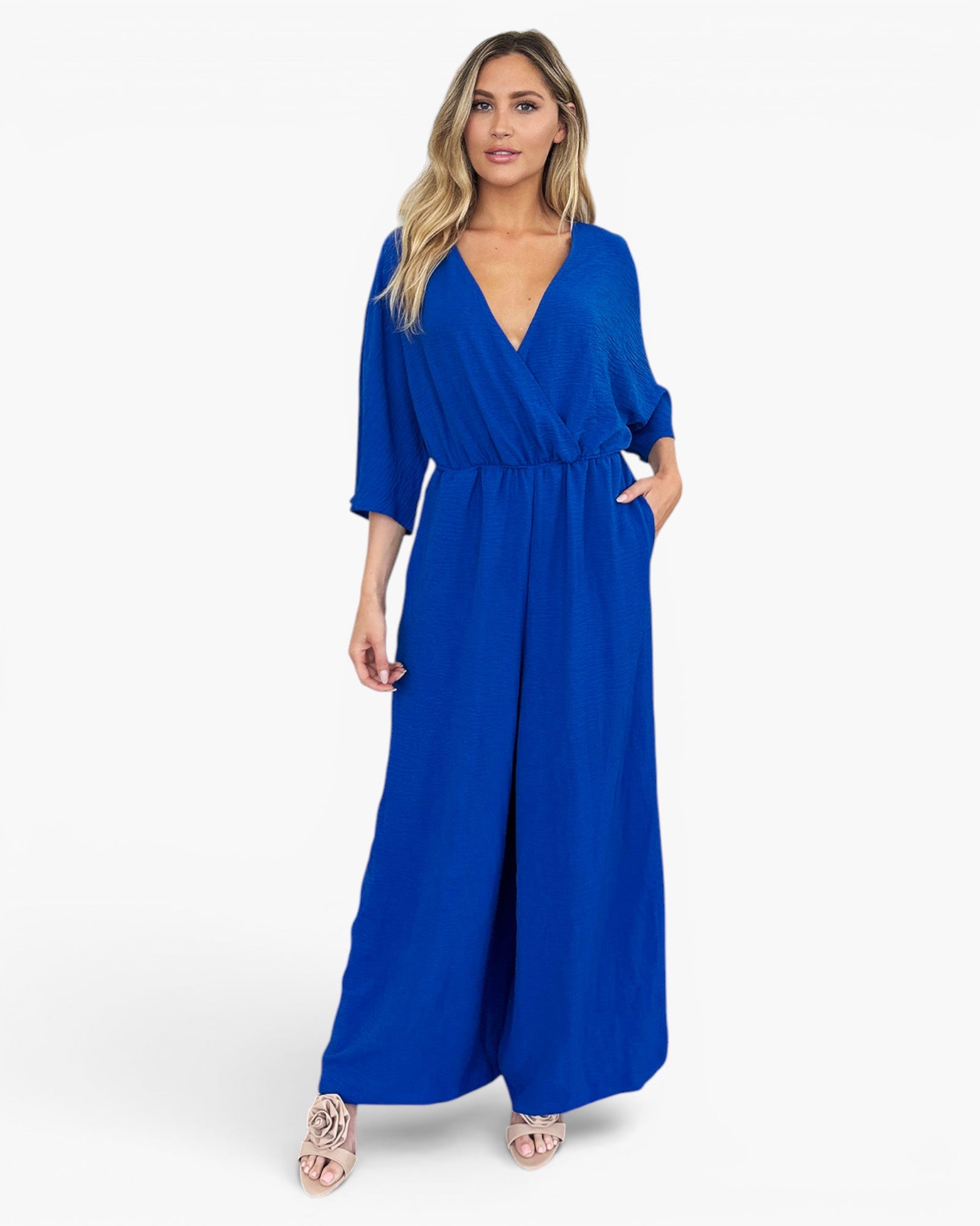 A woman modeling a royal blue jumpsuit with a wrap-style V-neck bodice, elbow-length sleeves, side pockets, and wide legs, photographed against a clean white studio background.
