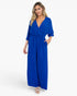 A woman modeling a royal blue jumpsuit with a wrap-style V-neck bodice, elbow-length flutter sleeves, side pockets, and a gathered waist, photographed against a clean white studio background.