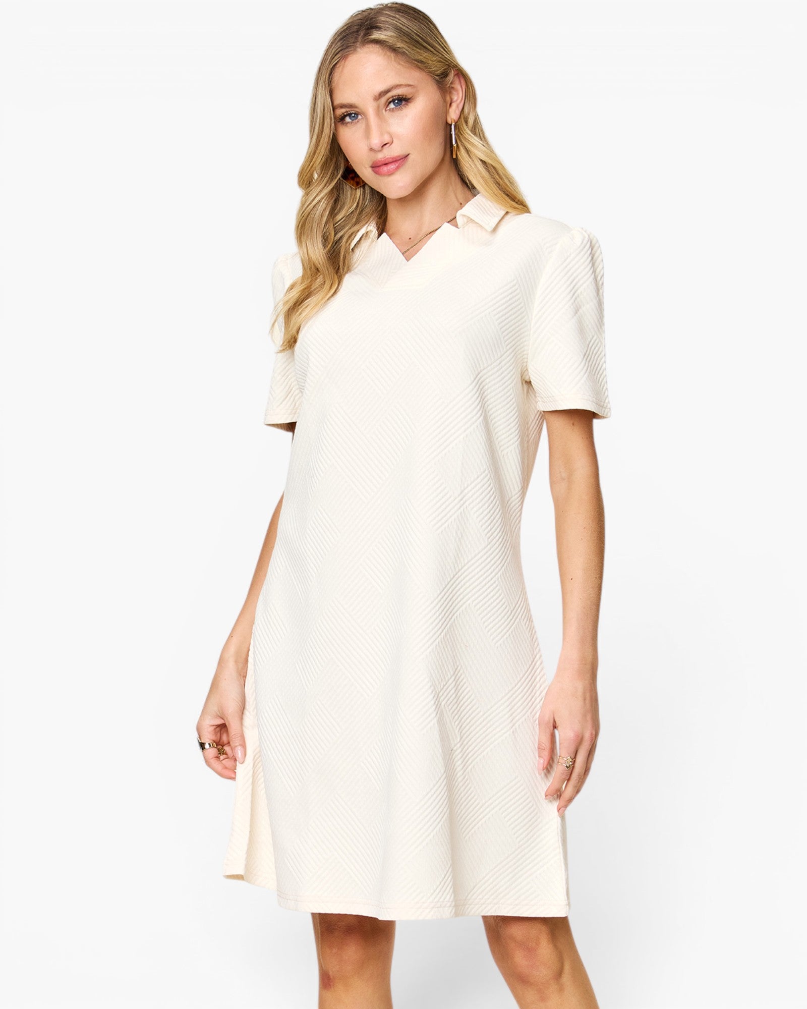 A woman modeling a short-sleeve collared shift dress in a warm off-white or cream color, featuring a subtle textured pattern, against a plain white studio background.