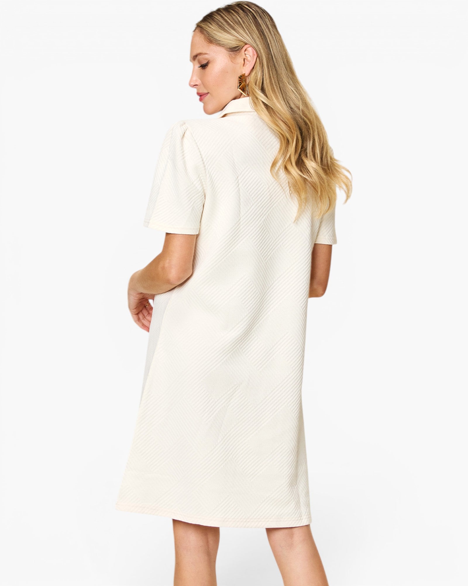 A woman with blonde hair is shown from the back wearing a short-sleeve, knee-length cream dress with a textured, waffle-like pattern. The background is a clean, solid white studio backdrop.
