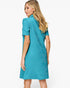 A woman modeling a short-sleeve teal dress with a textured geometric pattern, shown from a three-quarter back angle against a plain white studio background.