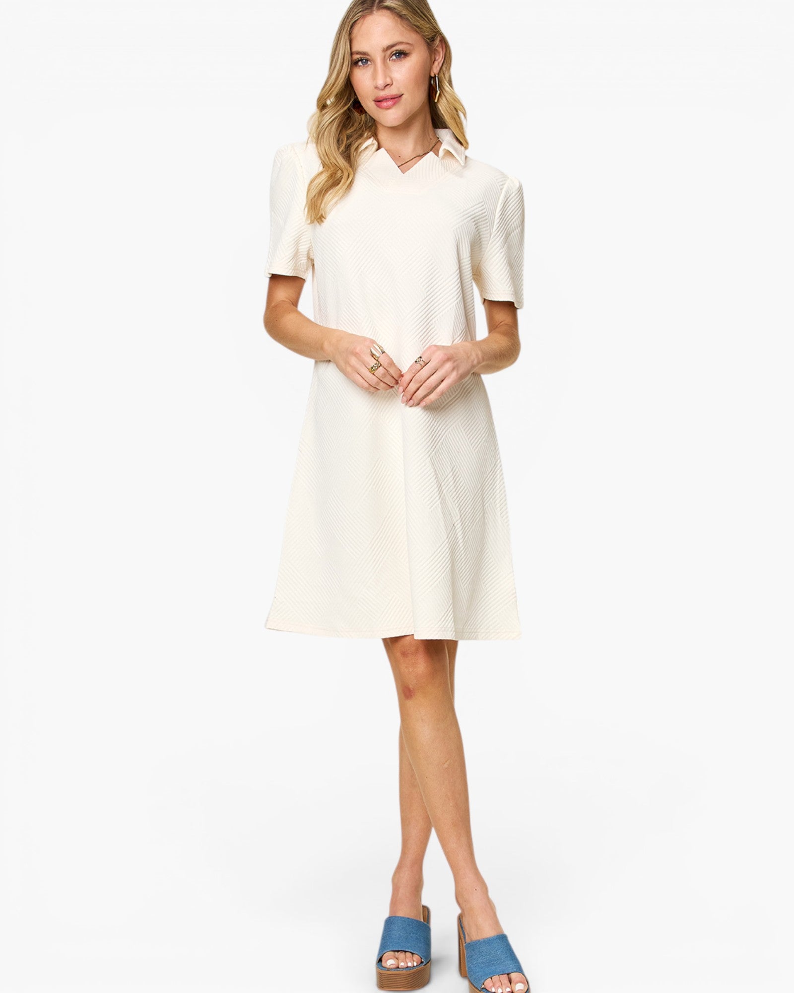A woman modeling a short-sleeve collared shift dress in off-white, paired with blue slide sandals, against a plain white studio background.