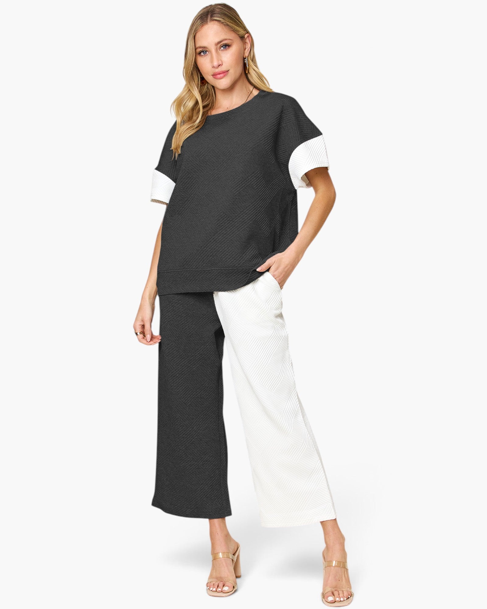A woman modeling a color-blocked short-sleeve top and matching wide-leg pants. The top is charcoal gray with white sleeve accents, and the pants are split down the middle with charcoal gray on one leg and white on the other.