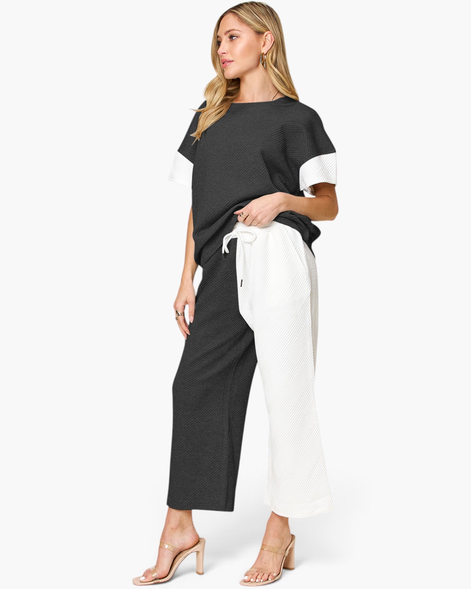 A woman modeling a two-tone color-blocked outfit consisting of a black short-sleeve top with white sleeve panels and black wide-leg cropped pants with white side panels, paired with clear-strap heeled sandals against a plain white background.