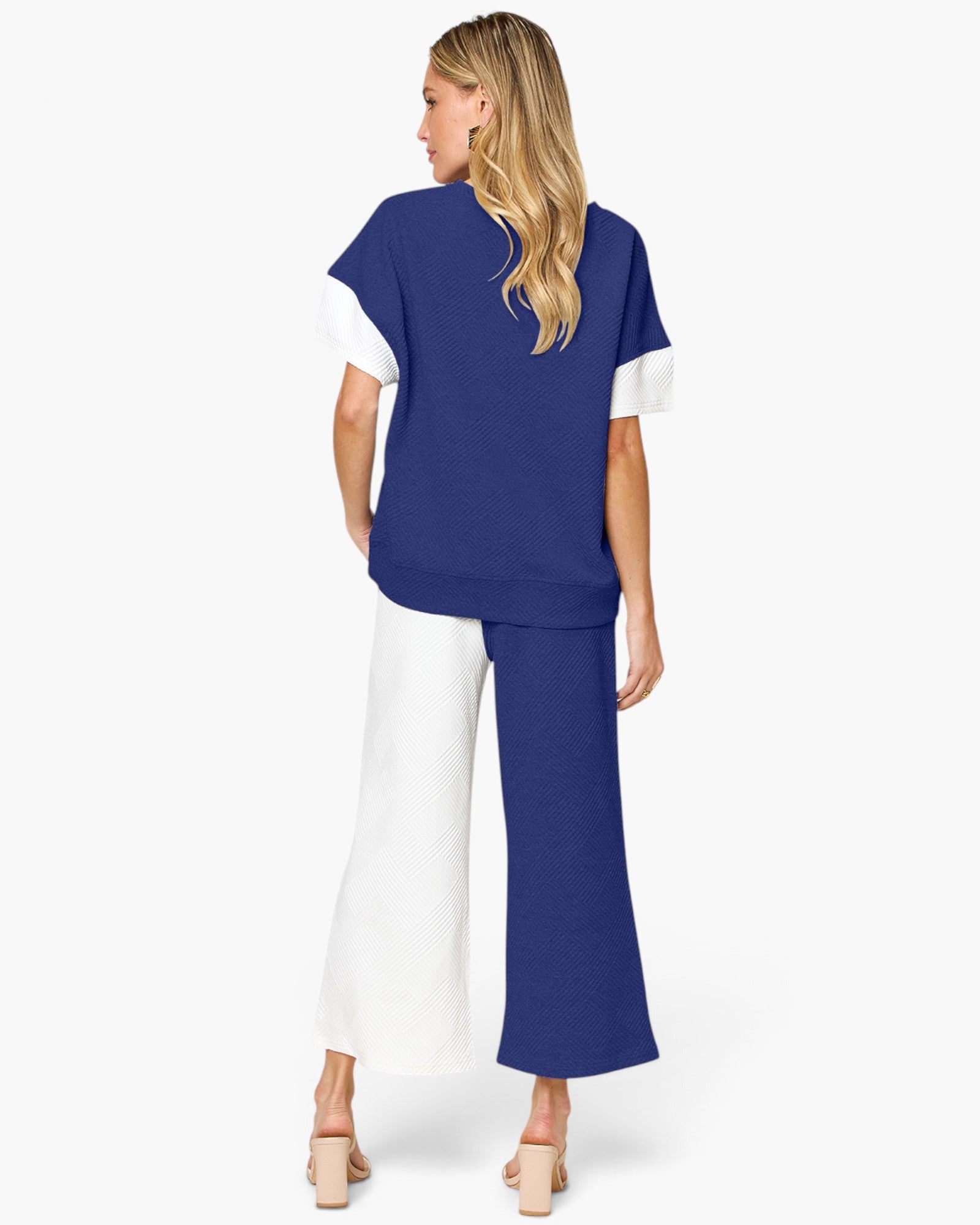 A woman shown from the back wearing a color-blocked knit top and matching wide-leg pants in royal blue and white, paired with beige block-heel sandals, against a light gray studio background.
