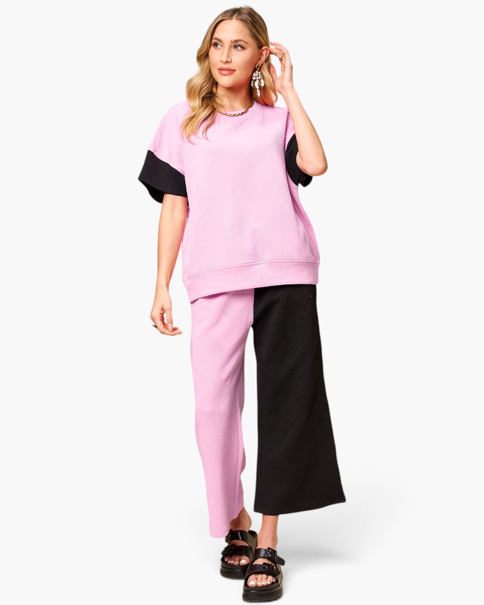 A woman modeling a color-blocked lounge set consisting of a short-sleeve sweatshirt and matching pants in pink and black, paired with black sandals against a light gray background.