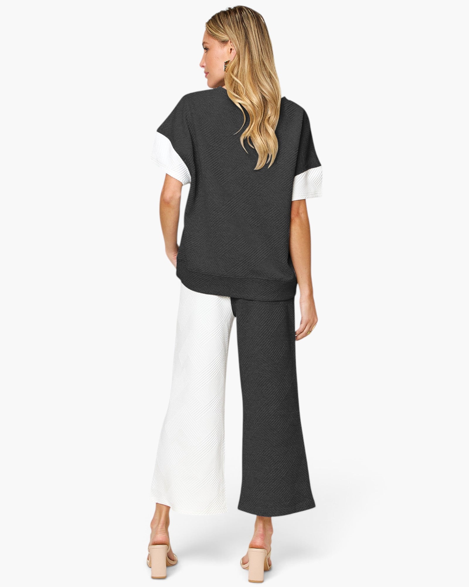A woman shown from the back wearing a color-blocked short-sleeve top and matching wide-leg pants in black and white, paired with beige high-heeled sandals, against a light gray studio background.
