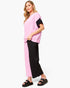 A woman modeling a color-blocked two-piece outfit consisting of a short-sleeve top with pink and black color blocking, and matching wide-leg pants in pink and black, paired with black slide sandals on a white studio background.