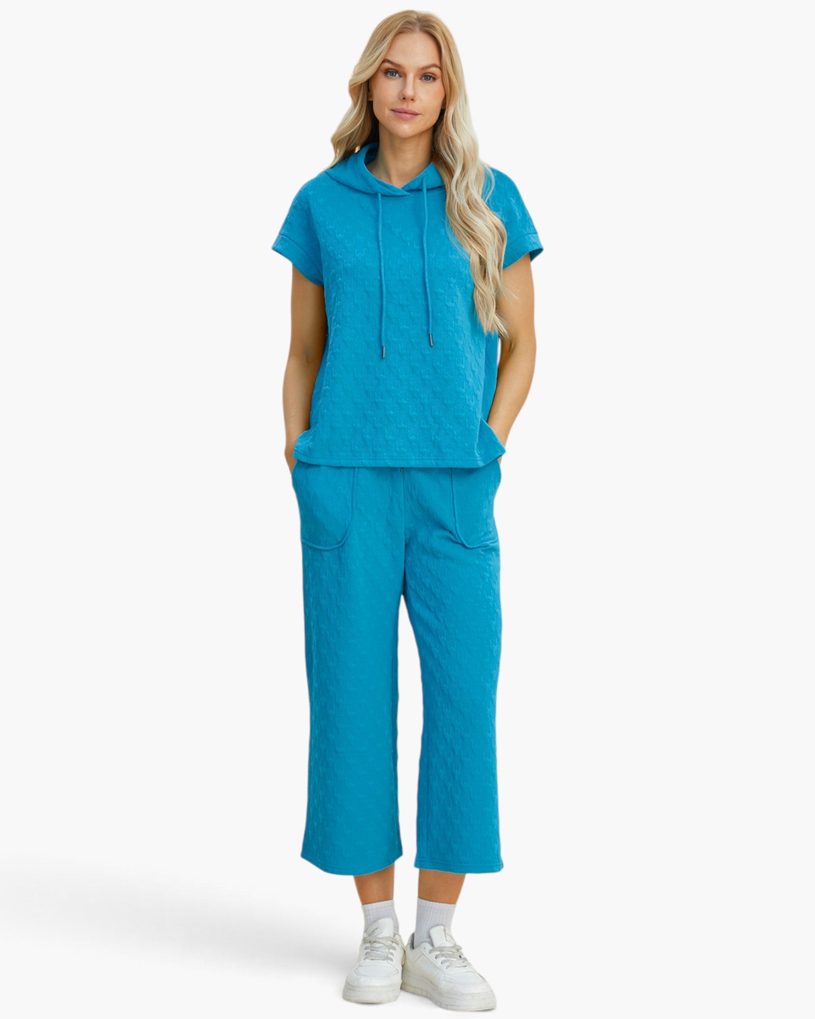 A woman modeling a matching bright turquoise blue loungewear set consisting of a short-sleeve cropped hoodie and wide-leg cropped pants, paired with white sneakers against a clean white studio background.