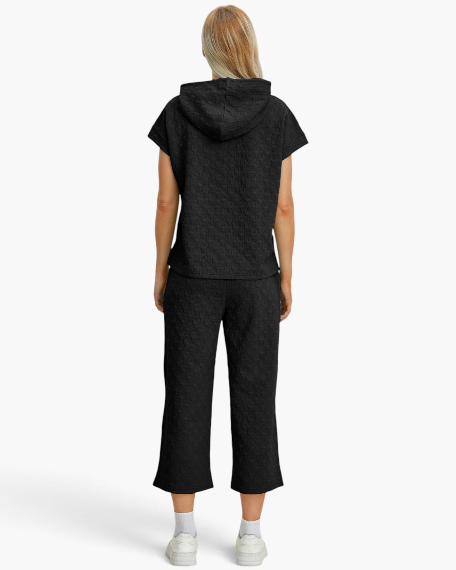 A model viewed from the back wearing a matching black textured hooded top and cropped pants set, paired with white socks and sneakers, against a plain white studio background.