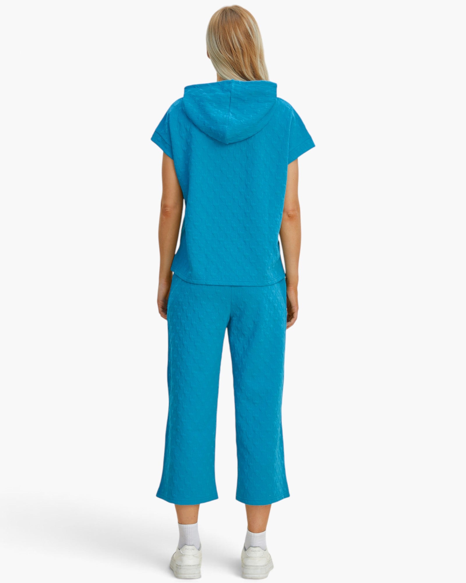 A model viewed from the back wearing a bright turquoise blue matching set consisting of a short-sleeve hooded top and cropped pants, paired with white socks and sneakers against a light gray studio background.