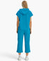 A model viewed from the back wearing a bright turquoise blue matching set consisting of a short-sleeve hooded top and cropped pants, paired with white socks and sneakers against a light gray studio background.