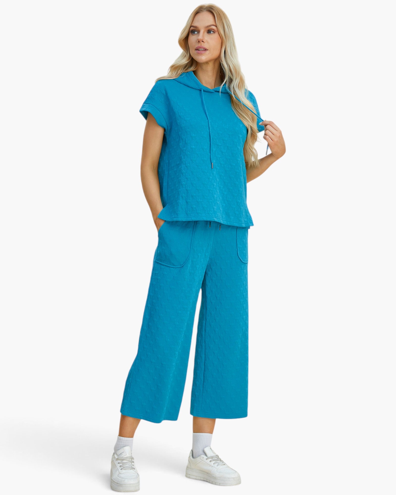 A woman modeling a bright turquoise blue matching lounge set consisting of a short-sleeve cropped hoodie top and wide-leg cropped pants, paired with white sneakers against a light gray studio background.