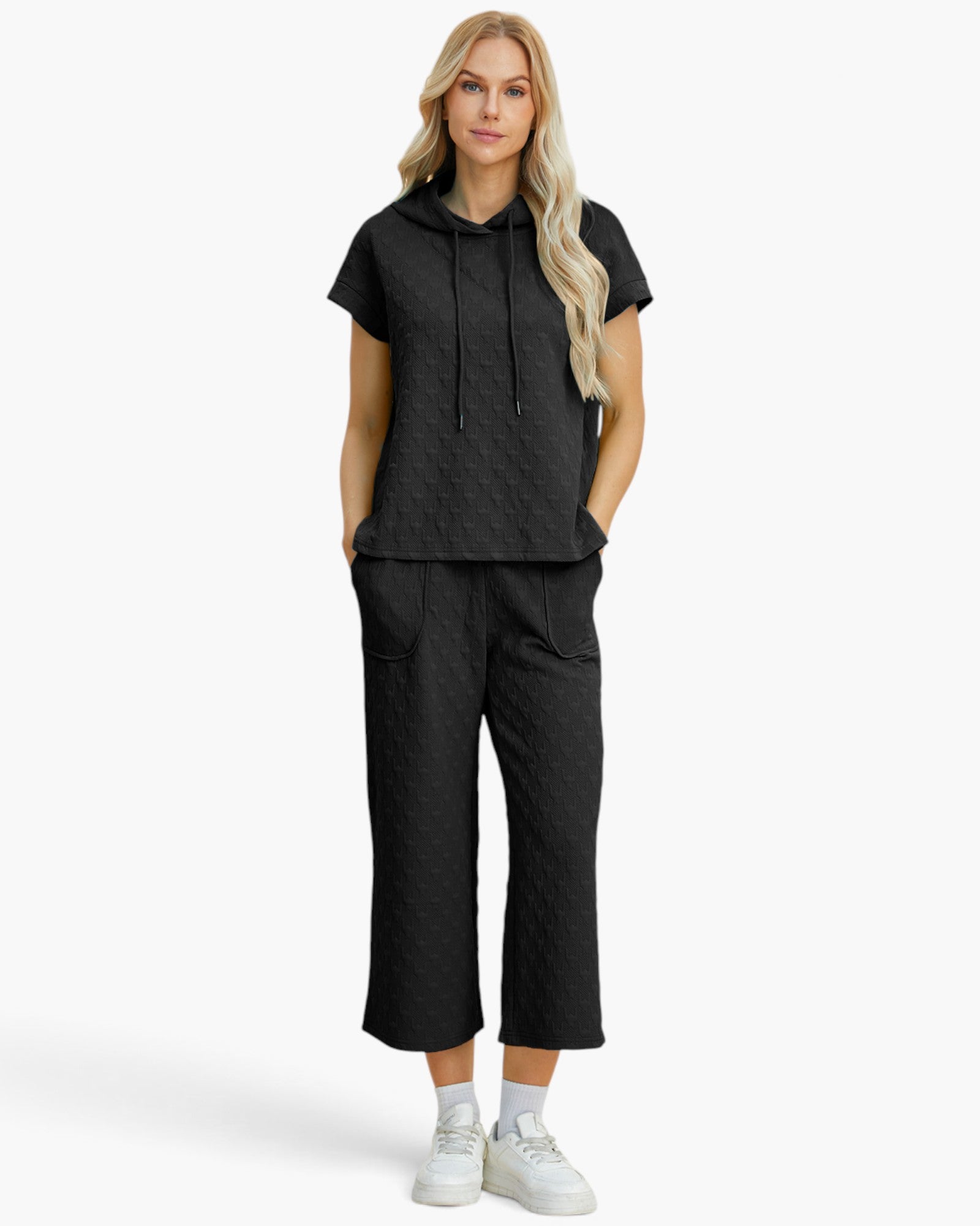 A woman modeling a matching black textured two-piece loungewear set consisting of a short-sleeve hooded top and cropped pants, paired with white sneakers against a light gray studio background.