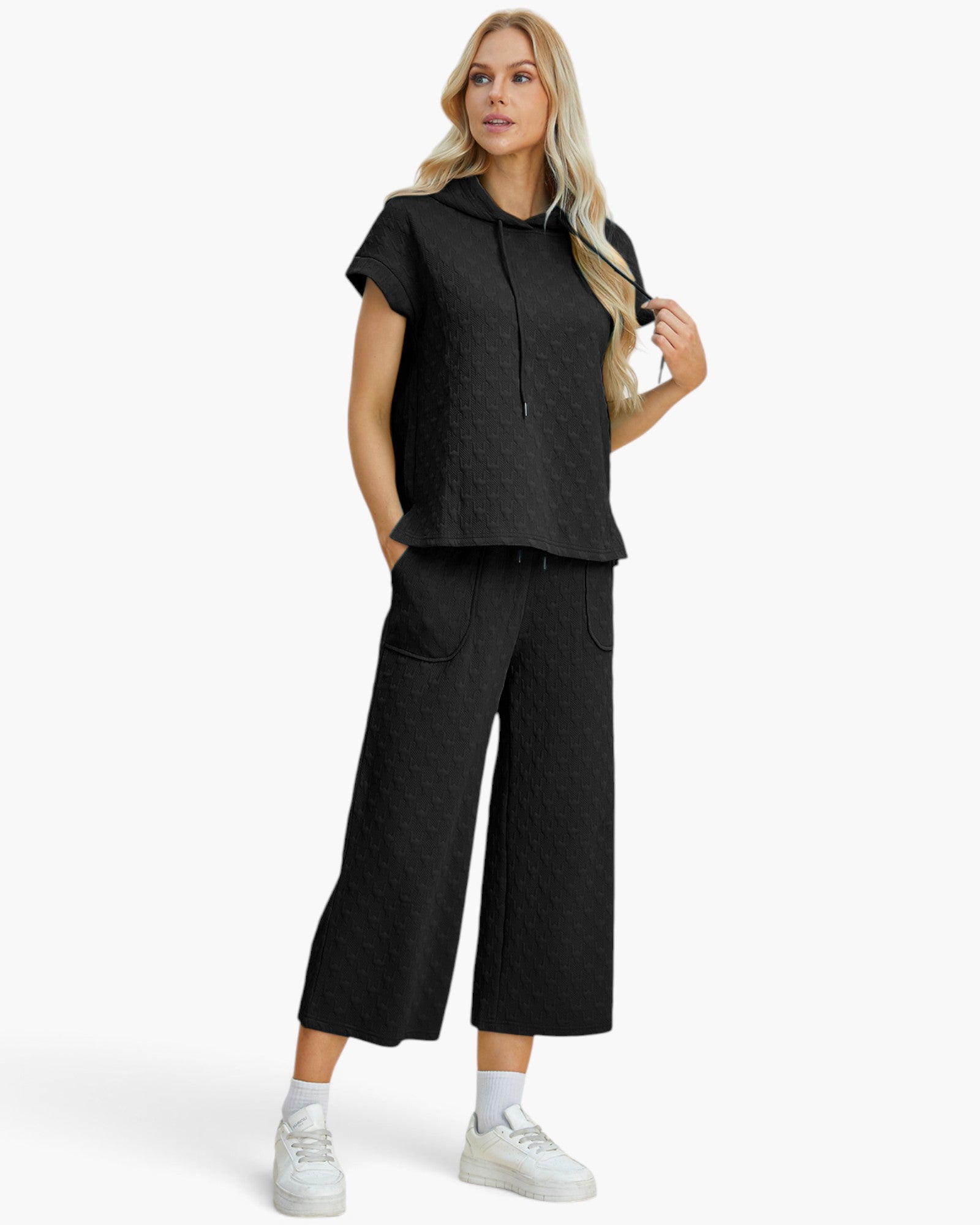 A woman modeling a matching black two-piece lounge set consisting of a short-sleeve textured hooded top and wide-leg cropped pants, paired with white sneakers against a light gray studio background.