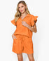 A woman modeling a bright orange two-piece set consisting of a short-sleeve collared top with ruffled sleeves and matching drawstring shorts, against a plain white studio background.