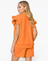 A woman shown from the back wearing a bright orange textured two-piece set consisting of a short-sleeve top with dramatic ruffled shoulder details and matching shorts, against a plain white studio background.