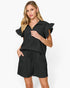 A woman modeling a black short-sleeve romper with ruffled flutter sleeves, a split V-neckline, and matching drawstring shorts against a plain white studio background.