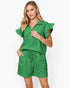 A woman modeling a bright green two-piece set consisting of a short-sleeve collared top with ruffled sleeves and matching drawstring shorts, against a plain white studio background.