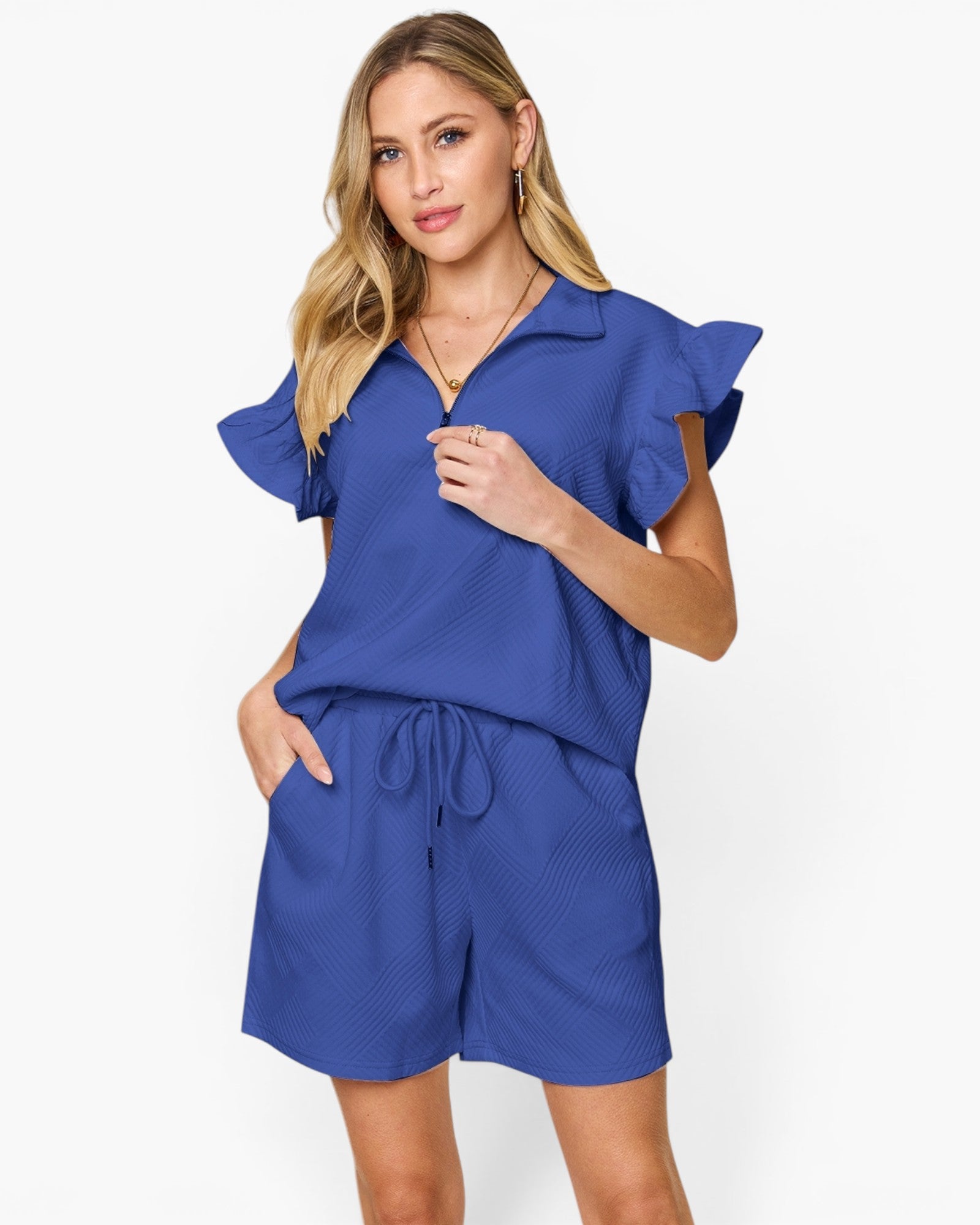 A woman modeling a royal blue two-piece romper set featuring a short-sleeve collared top with ruffled sleeves and matching drawstring shorts, against a plain white studio background.