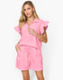 A woman modeling a bright pink two-piece set consisting of a short-sleeve collared top with ruffled sleeves and matching drawstring shorts, against a plain white studio background.