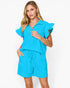 A woman modeling a bright turquoise blue short-sleeve romper with ruffled flutter sleeves and a drawstring waist, photographed against a clean white studio background.