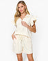 A woman modeling a cream-colored two-piece lounge set consisting of a short-sleeve top with ruffled sleeves and matching shorts with a drawstring waist, set against a plain white studio background.