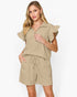 A woman modeling a light beige two-piece set consisting of a short-sleeve collared top with ruffled sleeves and matching drawstring shorts, against a plain white studio background.