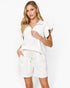 A woman modeling a white two-piece lounge set consisting of a short-sleeve collared top with ruffled sleeves and matching drawstring shorts, against a plain white studio background.
