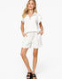 A woman modeling a white short-sleeve top with ruffled sleeves and matching white drawstring shorts, paired with black slide sandals, against a plain white studio background.