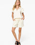A woman modeling a matching cream-colored two-piece set consisting of a short-sleeve ruffled collar top and drawstring shorts, paired with black slide sandals against a light gray background.