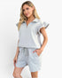 A woman modeling a light gray two-piece set featuring a short-sleeve collared top with a half-zip and ruffle sleeves, paired with matching drawstring shorts, against a plain white studio background.