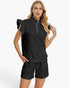 A woman modeling a black textured two-piece lounge set consisting of a short-sleeve quarter-zip top with ruffled sleeves and matching drawstring shorts, against a plain white studio background.