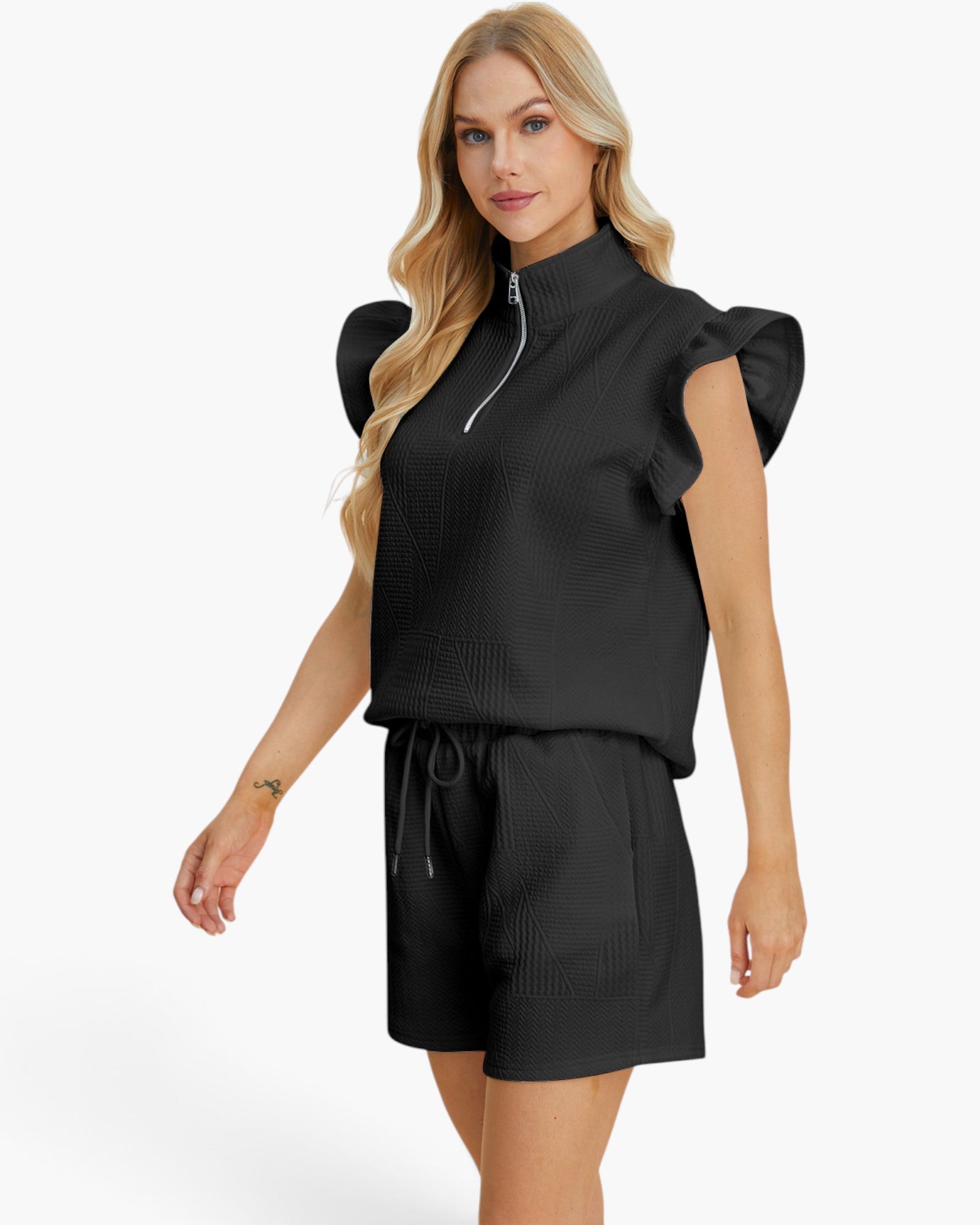 A blonde model wearing a black ribbed knit two-piece set consisting of a mock-neck short-sleeve top with a half-zip and ruffled sleeves, paired with matching drawstring shorts, against a plain white background.