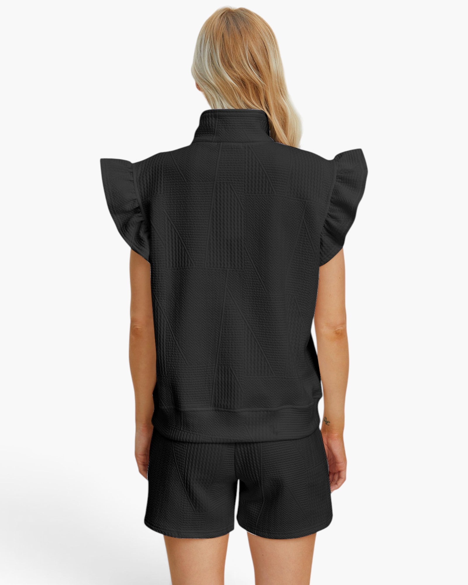 A woman shown from the back wearing a black sleeveless top with dramatic ruffled shoulders and matching black textured shorts against a plain light background.