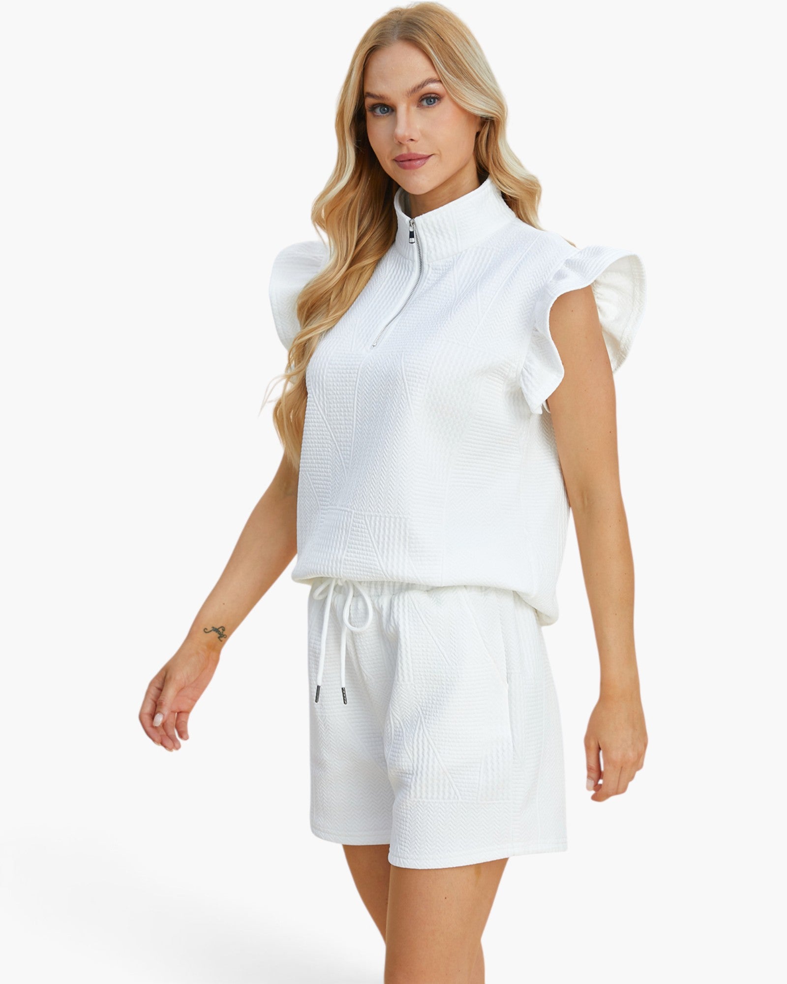 A blonde model wearing a white textured two-piece lounge set consisting of a sleeveless mock-neck top with ruffle details and matching drawstring shorts, set against a plain white background.