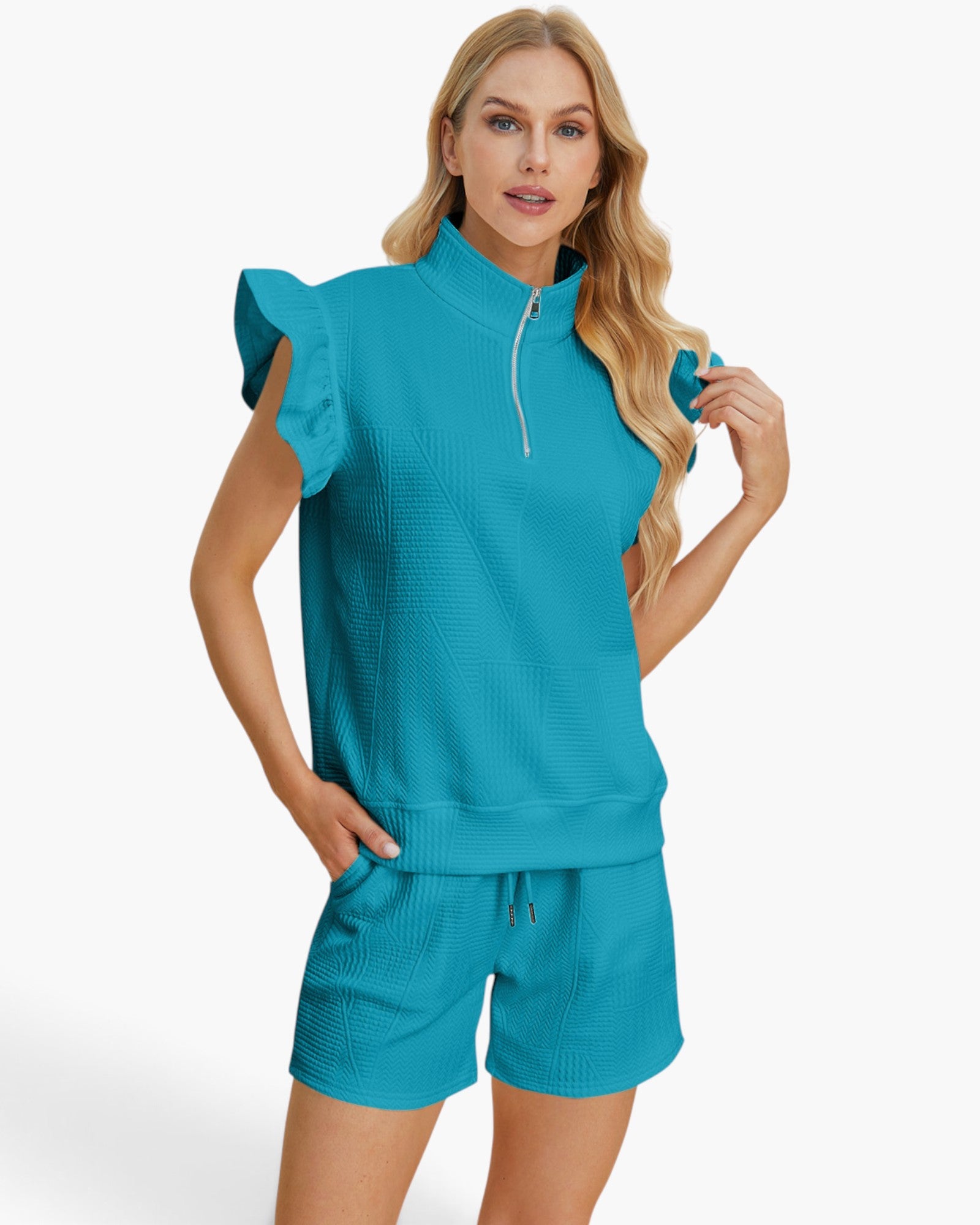 A woman modeling a matching turquoise blue lounge set consisting of a sleeveless quarter-zip top with ruffle details and matching drawstring shorts, set against a plain white studio background.