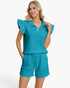 A woman modeling a matching two-piece lounge set in a bright turquoise blue-green color. The set includes a short-sleeve top with ruffled cap sleeves and a half-zip collar, paired with matching textured drawstring shorts, against a clean white studio background.